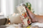 Preview: Thermobecher mit kunstvollem Violin-Design, kombiniert mit floralen Mustern und Musiknoten, gehalten in einer Hand vor einem gemütlich beleuchteten Innenraum mit unscharfen, warmen Lichtakzenten im Hintergrund.